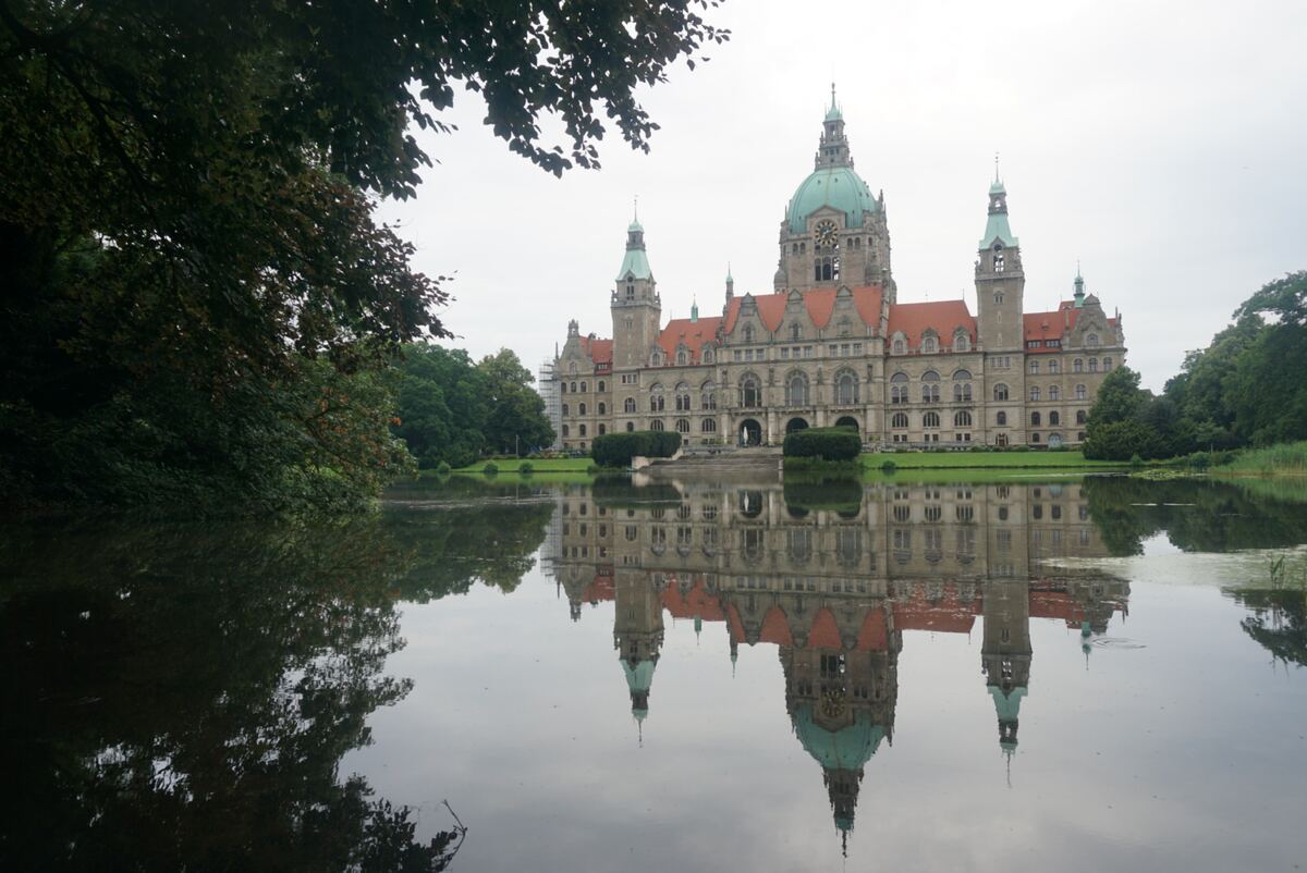 Een breed shot van het Nieuwe Raadhuis in Hannover, Duitsland, weerspiegeld in het water van de vijver ervoor. Het gebouw heeft een grote koepel met een klok en meerdere torens met groene daken. Rondom het water is groen gras en aan de zijkanten staan bomen en struiken. De lucht is bewolkt.