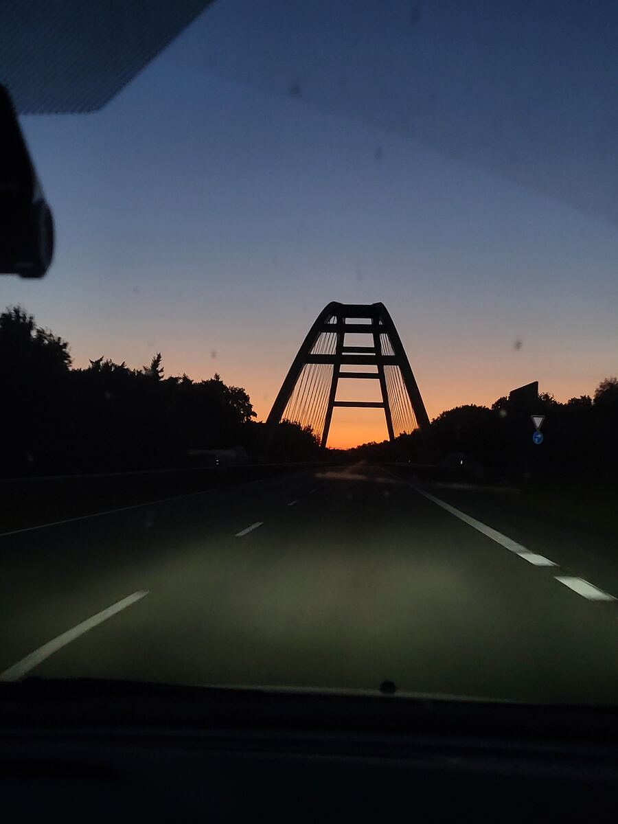 Een foto genomen vanuit een auto, rijdend op een snelweg in de schemering. De koplampen verlichten de weg. In de verte is de silhouet van een grote tuibrug zichtbaar tegen een oranje-blauwe avondlucht. Aan weerszijden van de weg staan donkere bomen. Rechts is een verkeersbord te zien.