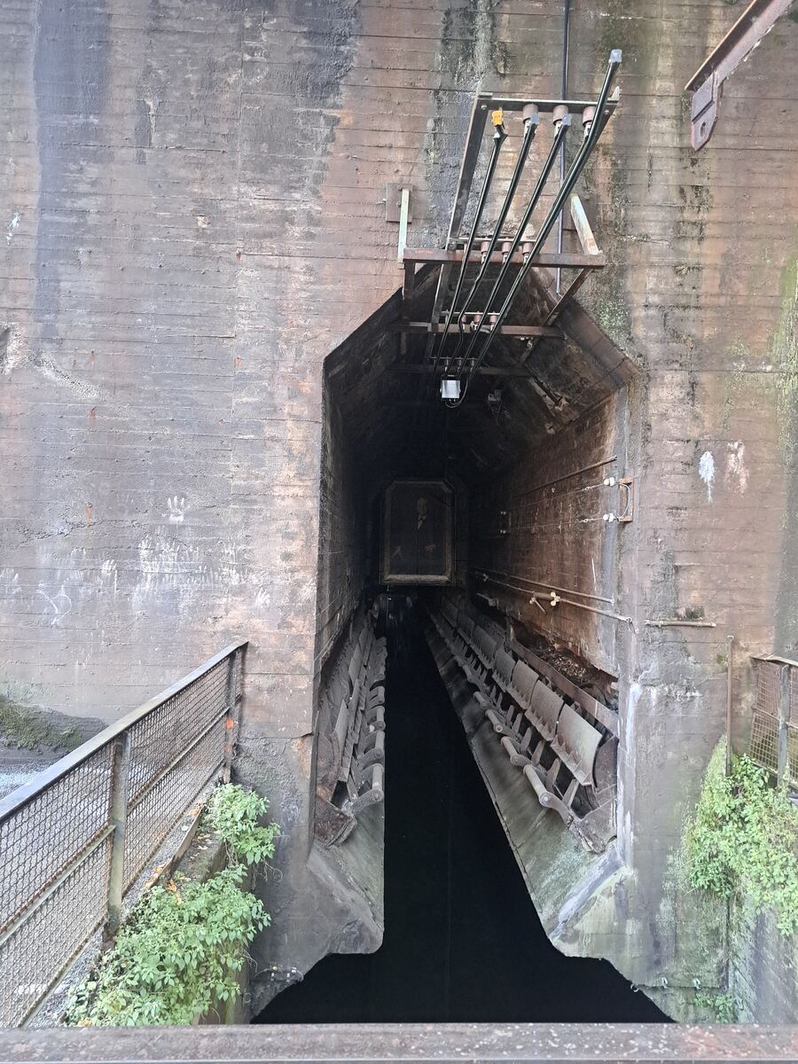 De foto toont een donkere, diepe schacht of tunnelopening, omgeven door robuuste betonnen muren. Aan de zijkanten van de tunnel zijn metalen constructies en wat lijkt op transportbanden of goten zichtbaar, met daarin donker water of vloeistof. Boven de opening hangen industriële leidingen of kabels. Op de voorgrond is een deel van een metalen hekwerk met klimop te zien. Diep in de tunnel hangt een ingelijst portret van een man.