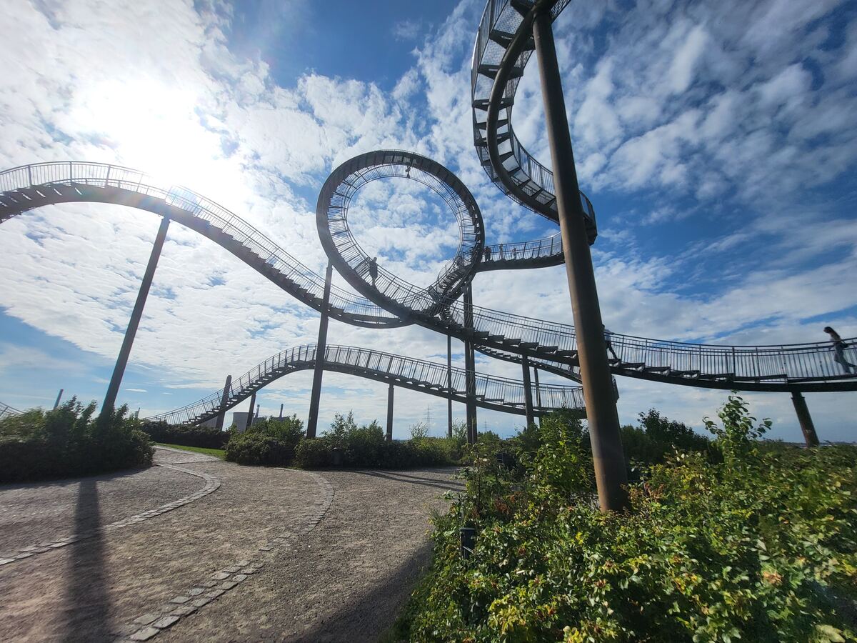 Een sculptuur van staal, de 'Tiger & Turtle – Magic Mountain', met trappen die een achtbaan nabootsen, staat onder een helderblauwe lucht met witte wolken. De zon schijnt fel en er zijn enkele mensen te zien die over de constructie lopen. Op de voorgrond is een grindpad en groen struikgewas zichtbaar.