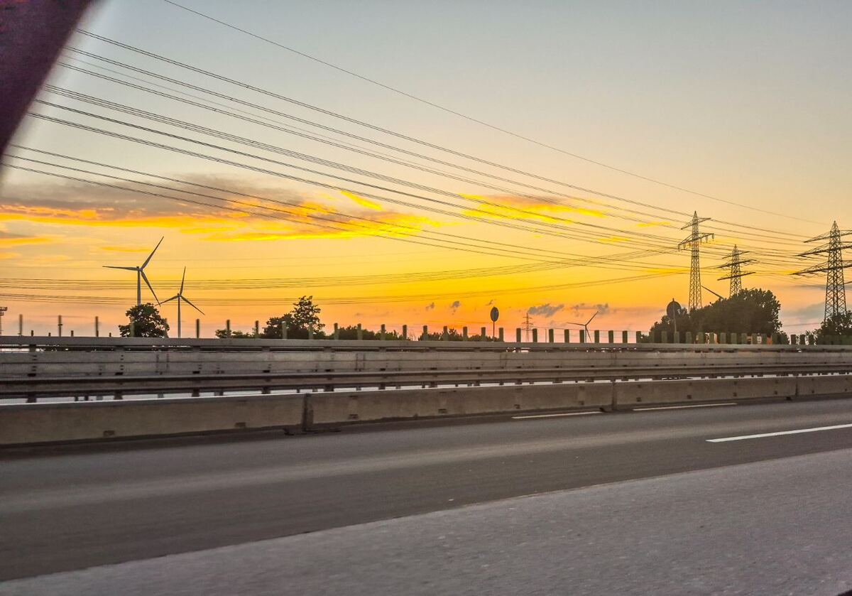 Een foto genomen vanuit een rijdende auto, met op de voorgrond een snelweg en vangrails. Op de achtergrond is een zonsondergang te zien met een oranjegele lucht. Er staan windmolens en hoogspanningsmasten langs de weg.