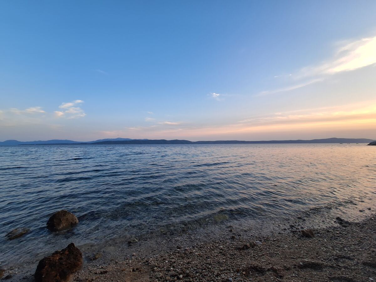 Een weids uitzicht over een kalme zee met een kiezelstrand op de voorgrond. De horizon toont een reeks bergen onder een lucht die van blauw naar zachtroze en oranje kleurt, wat duidt op zonsondergang. Enkele rotsen liggen in het ondiepe water.