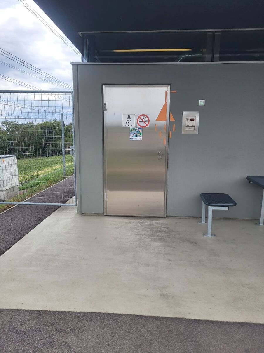 Een roestvrijstalen deur van een modern gebouw met een douche- en rookverbodsymbool, naast een betaalautomaat en een bankje. Op de achtergrond is een hekwerk en groen landschap met elektriciteitsmasten te zien onder een bewolkte lucht.