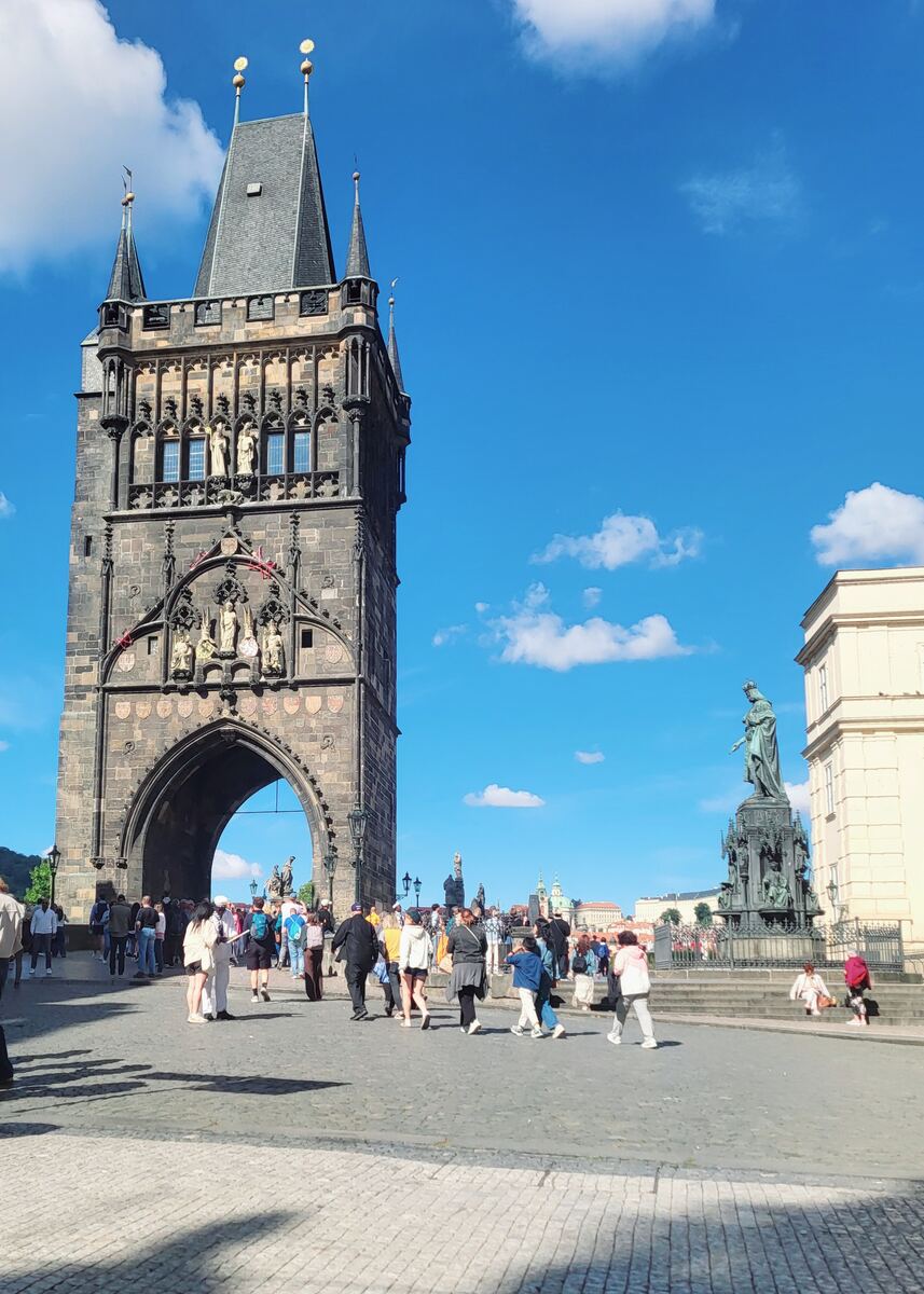Een foto van de Karelsbrugtoren in Praag, met veel mensen op de geplaveide straat ervoor. De toren is een imposant gotisch bouwwerk met een grote boog en gedetailleerde sculpturen. Rechts van de toren staat een groot standbeeld op een voetstuk. De lucht is helderblauw met enkele witte wolken.