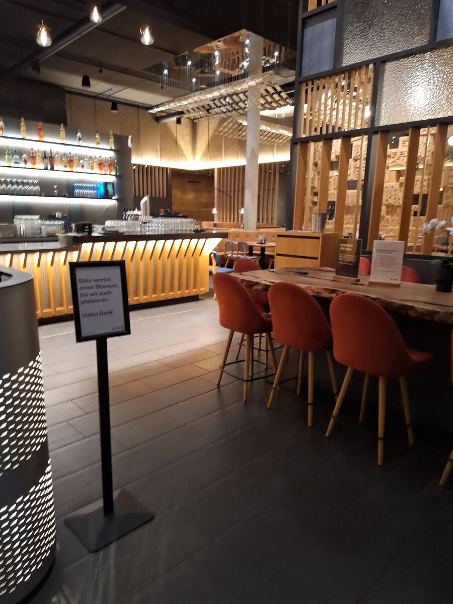 Een interieurfoto van een modern restaurant of bar met een lange bar aan de linkerkant, verlichte schappen met flessen, en een rij oranje stoelen aan een houten tafel aan de rechterkant. Een bordje met Duitse tekst staat op de voorgrond.