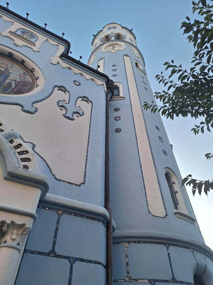 Een opvallende blauw-witte kerk met een hoge toren en een mozaïek boven de ingang, gefotografeerd vanuit een laag perspectief tegen een heldere lucht. De kerk heeft decoratieve elementen en een klok in de toren.