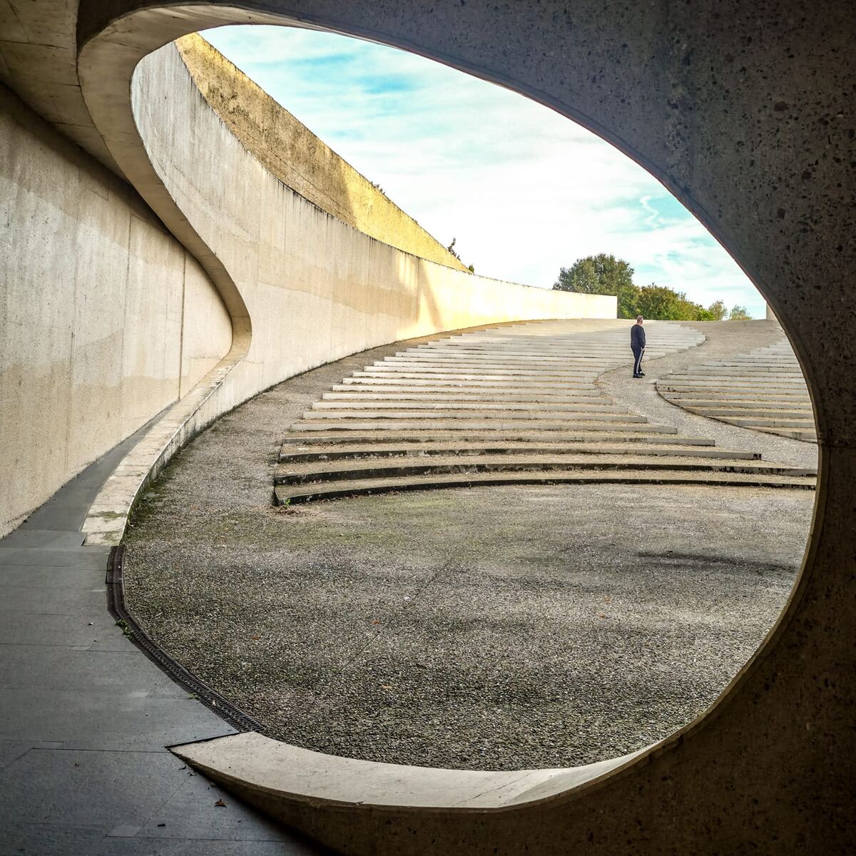 Een persoon staat op een brede trap van een modern amfitheater, omringd door gebogen betonnen muren. De zon schijnt op de bovenrand van de muur en de lucht is deels bewolkt.