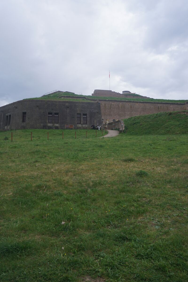 Een groot, oud fort met een stenen muur en een met gras begroeide wal. Op de top van de wal wappert een rood-wit-blauwe vlag aan een paal. Een pad van grind leidt omhoog naar een ingang in de muur, waar enkele mensen in de verte bij staan. De voorgrond bestaat uit een groen grasveld onder een bewolkte lucht.