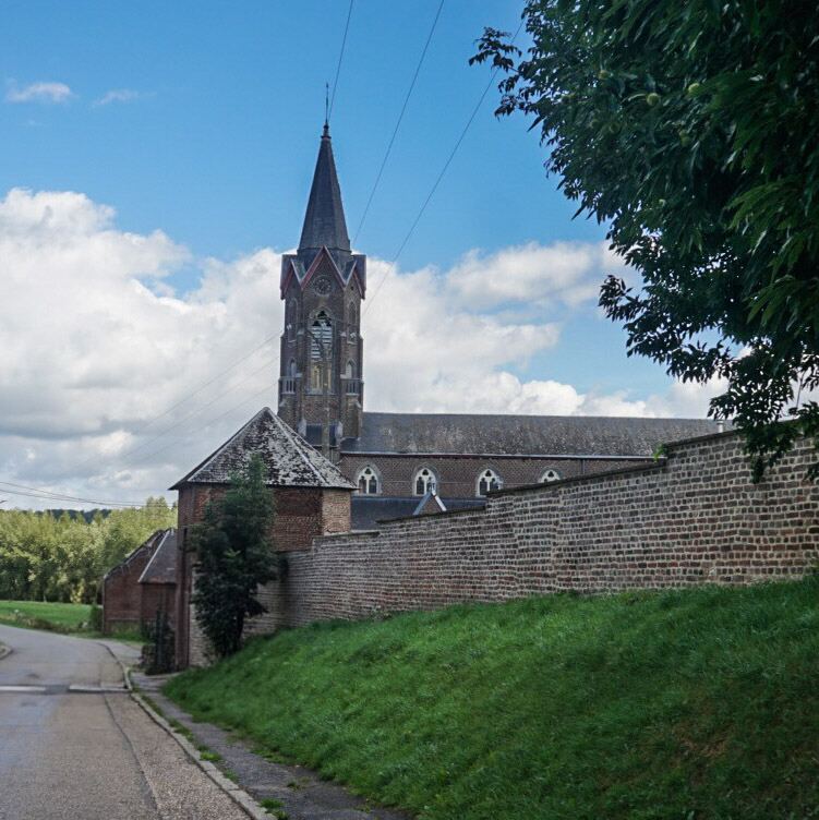 Een foto van een kerk met een hoge, spitse toren en een bakstenen muur op de voorgrond. De kerk heeft gotische ramen en een donker dak. Links van de muur loopt een geasfalteerde weg en is een groene berm te zien. De lucht is blauw met witte wolken.