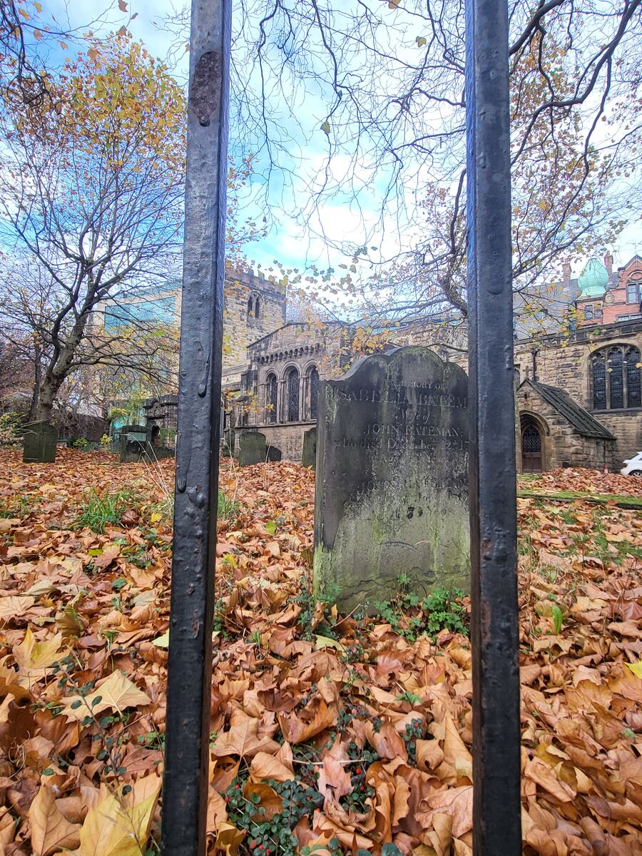 Een herfstige begraafplaats met een oude grafsteen op de voorgrond, gezien door de spijlen van een hek. Op de achtergrond staat een stenen kerkgebouw met een toren en rondboogvensters. De grond is bedekt met bruine en gele bladeren, en de bomen hebben nog enkele gele bladeren.