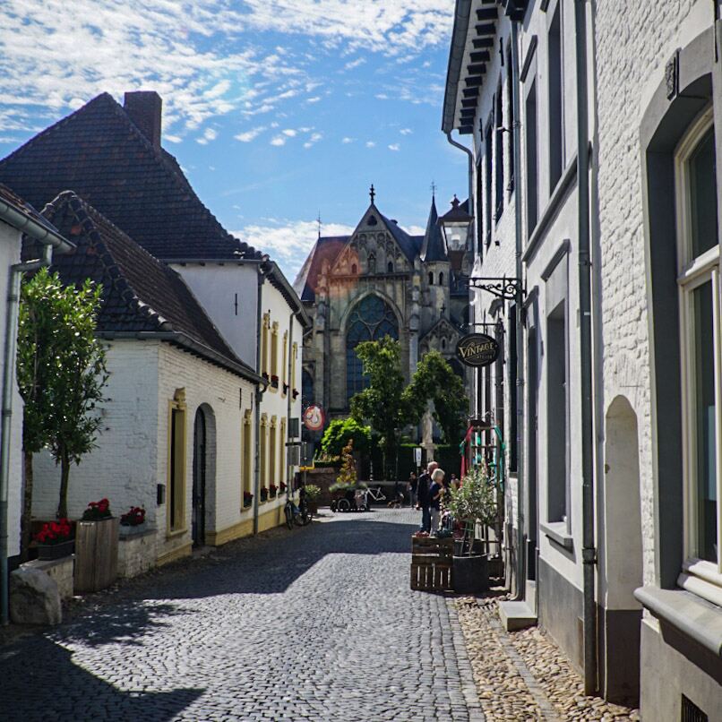 Een smalle kasseienstraat met aan weerszijden witte huizen en op de achtergrond een grote kerk met gotische architectuur. De lucht is blauw met verspreide wolken.