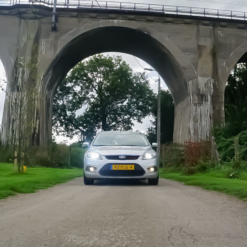 Een zilverkleurige Ford Focus station staat op een onverharde weg onder een groot stenen viaduct met meerdere bogen. Op de achtergrond is groen gebladerte van bomen te zien en een bewolkte lucht. De koplampen van de auto zijn aan.