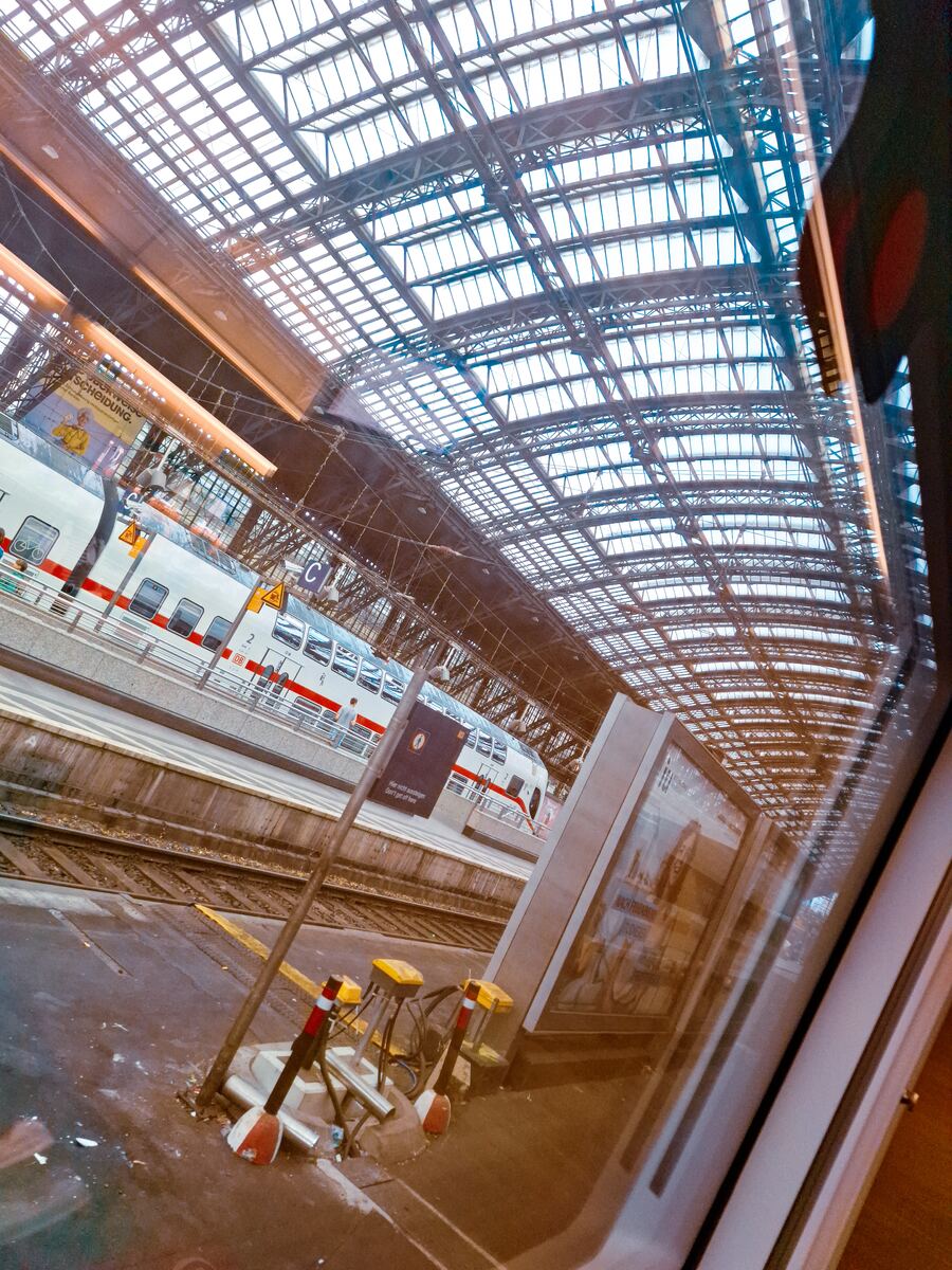 Een schuin genomen foto van een treinstation met een groot glazen dak. Op de sporen staat een witte trein met rode strepen en het DB-logo. Er zijn perrons zichtbaar en een reclamebord.