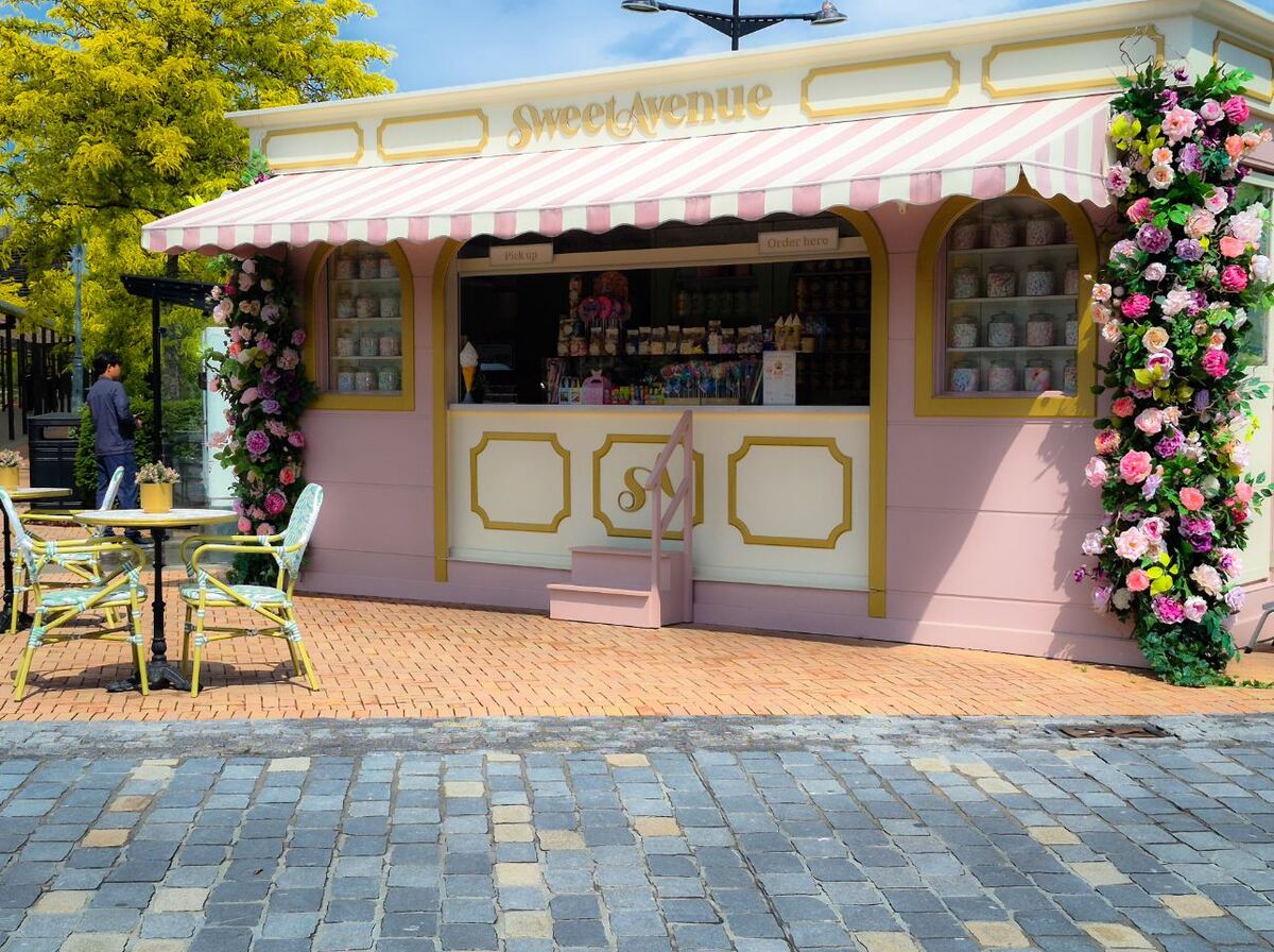 Een pastelroze kraam met de naam 'Sweet Avenue' en een gestreepte luifel, versierd met kleurrijke bloemen. Voor de kraam staan twee tafels met stoelen op een terras van klinkers, en op de achtergrond is een boom met geelgroene bladeren te zien. Een persoon staat links van de kraam.