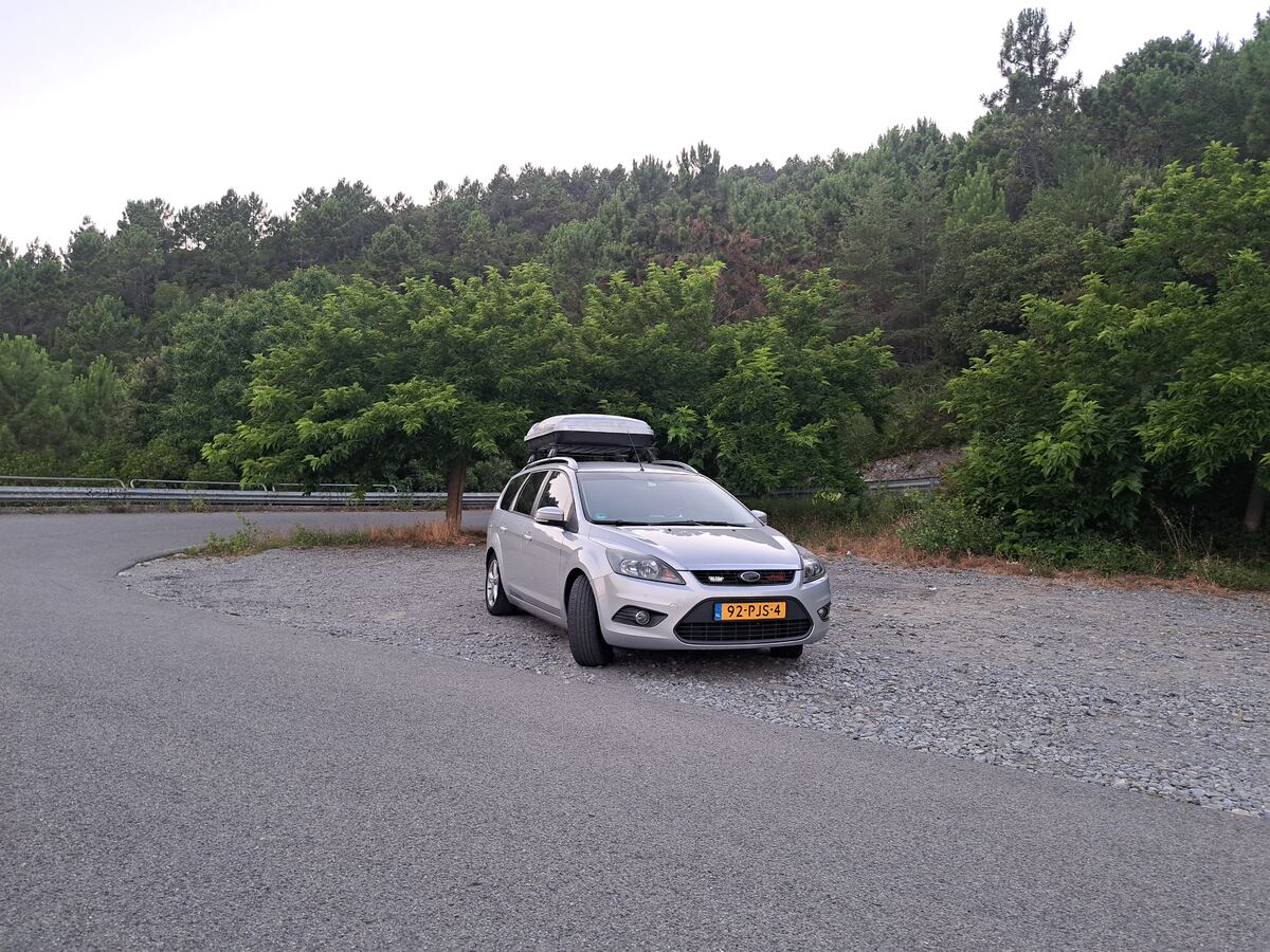 Een zilverkleurige Ford Focus stationwagen, voorzien van een dakkoffer, staat geparkeerd op een onverharde strook naast een geasfalteerde weg. De auto heeft een Nederlands kenteken '92-PJS-4'. Op de achtergrond is een dichtbegroeide heuvel of bergflank te zien met voornamelijk groene bomen, waaronder zowel loof- als naaldbomen. Langs de weg loopt een metalen vangrail. De lucht boven de heuvel is licht en egaal, wat duidt op schemering.
