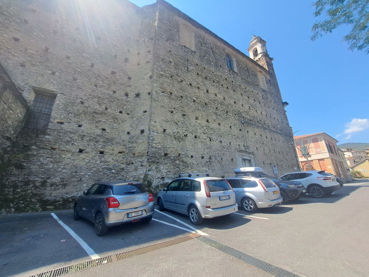 Een imposante, hoge stenen muur van een oud gebouw domineert de linkerzijde van de foto, met daarlangs geparkeerde auto's. De muur is opgebouwd uit onregelmatige stenen en heeft meerdere kleine, vierkante openingen en enkele ramen. Boven de muur is een deel van een klokkentoren zichtbaar tegen een helderblauwe lucht. Rechts op de achtergrond zijn andere gebouwen met terracotta daken en heuvels te zien. De zon schijnt fel, wat strepen van licht veroorzaakt in de bovenhoek van de foto. Op de voorg
