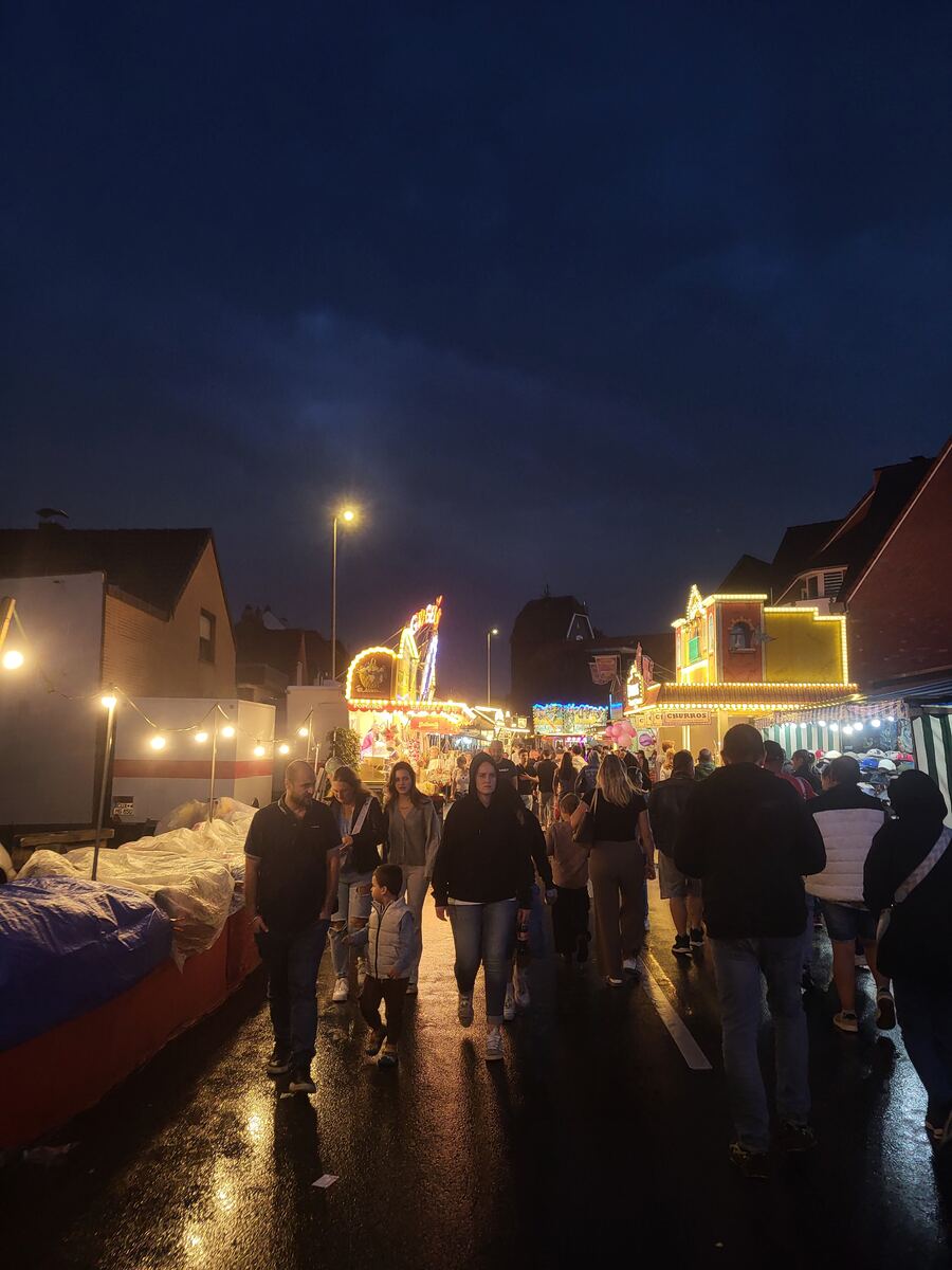 Een avondfoto van een drukke kermisstraat, verlicht door kermislampen en straatverlichting. Mensen lopen over de natte straat, die het licht reflecteert. Op de achtergrond zijn kermisattracties en kraampjes te zien, met woonhuizen aan de zijkanten.