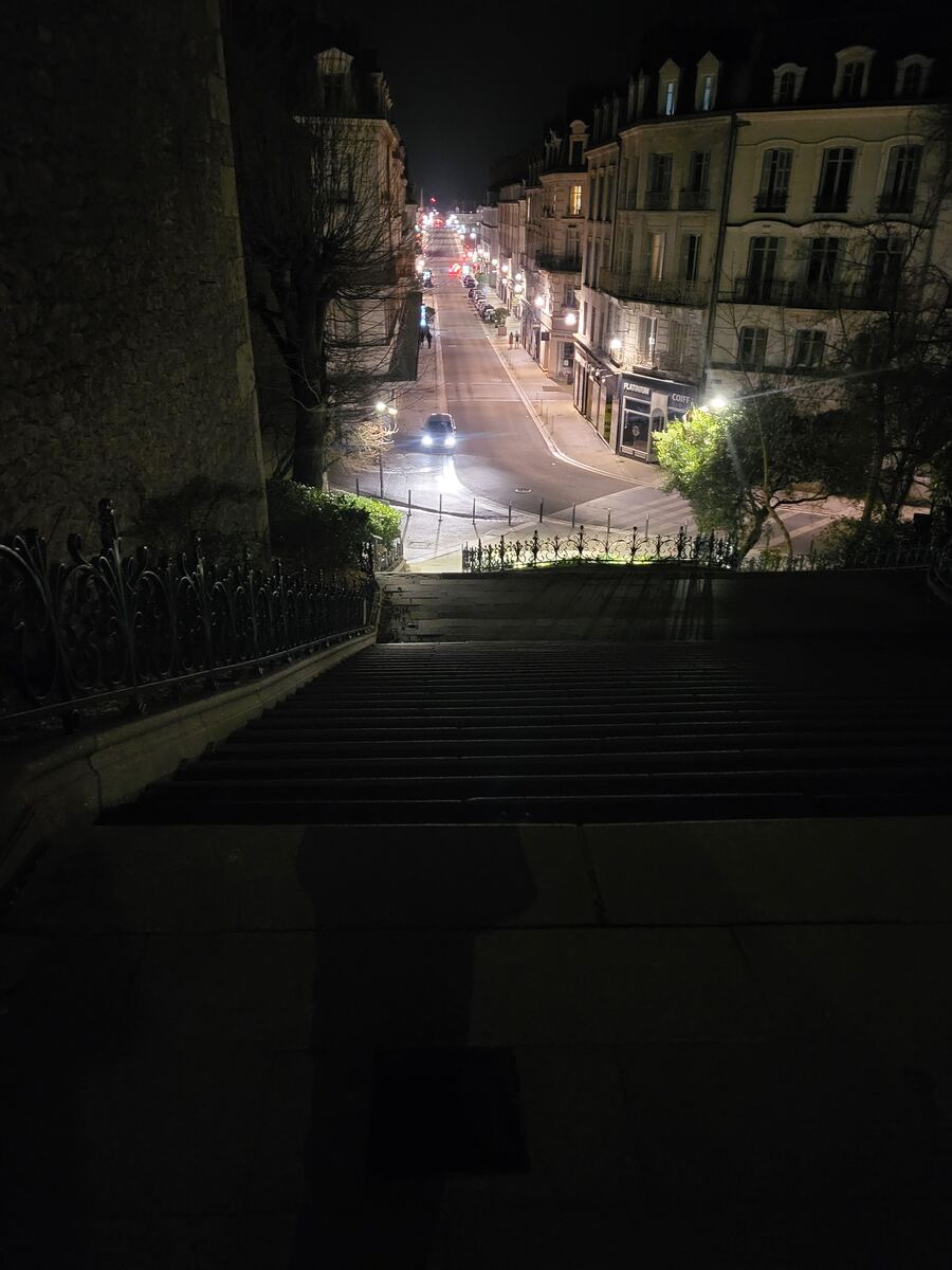 Een nachtelijke foto genomen vanaf een hoge trap die uitkijkt op een verlichte straat met gebouwen aan weerszijden. Er rijdt een auto met koplampen aan over de weg. Links is een hoge stenen muur zichtbaar en onderaan de trap is een decoratief hekwerk.