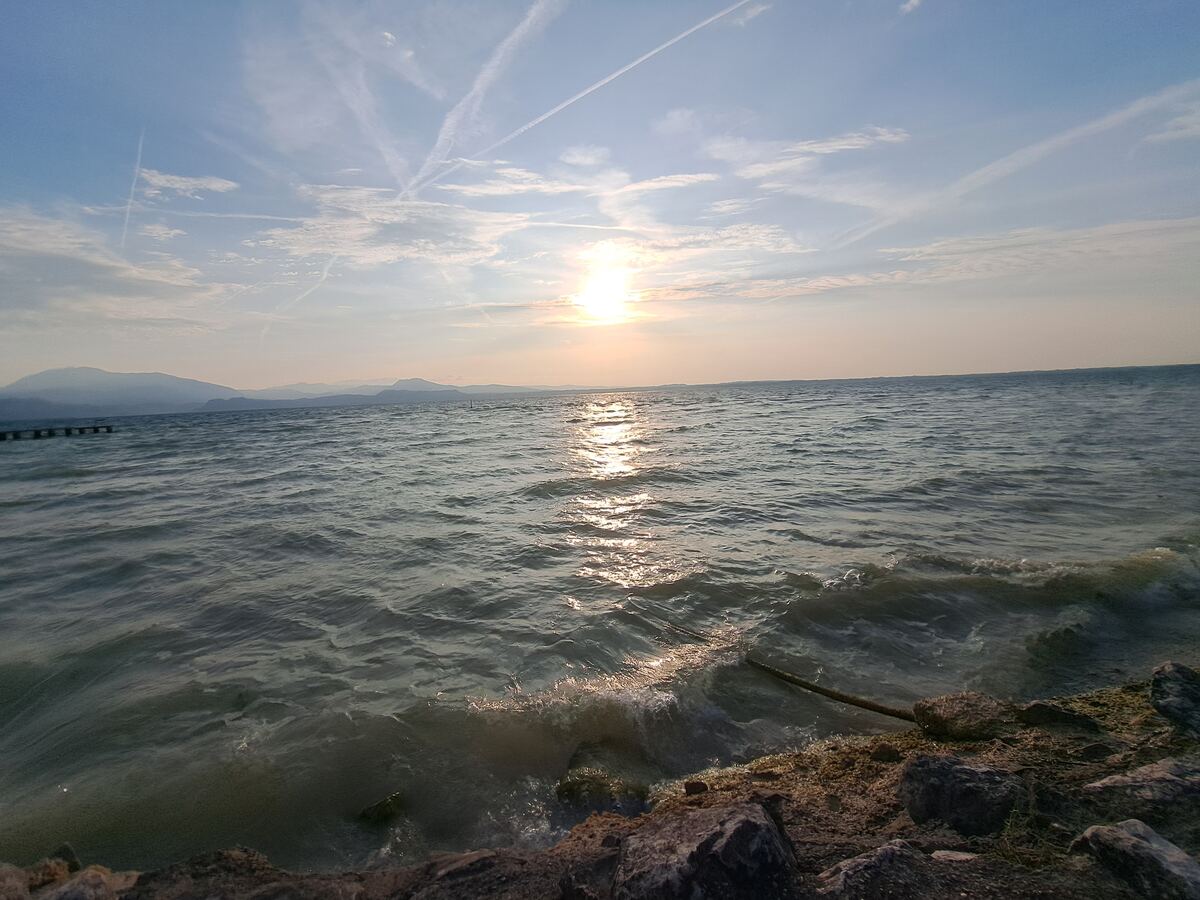 De foto toont een zonsopkomst boven een groot meer, met de zon laag aan de horizon die reflecteert op het water. Aan de linkerkant zijn bergen zichtbaar in de verte en een houten steiger. De voorgrond bestaat uit rotsen en golvend water.
