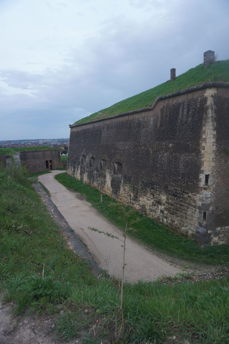 Een onverhard pad loopt langs een hoge, stenen vestingmuur die deels begroeid is met gras. Aan de linkerkant is een deel van een andere vestingmuur zichtbaar met een open doorgang. Op de achtergrond is een stad te zien onder een bewolkte lucht.