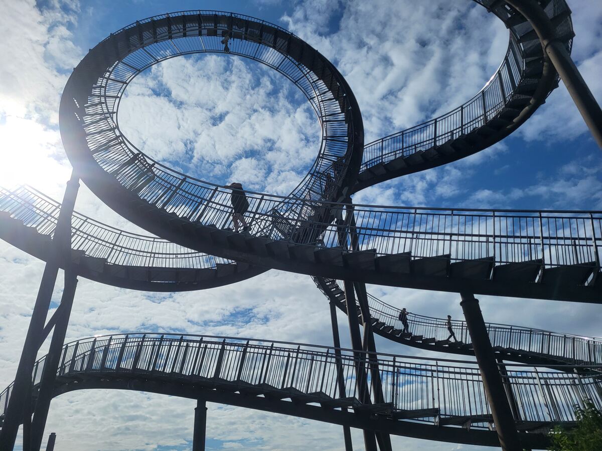 Een foto genomen vanuit een laag standpunt van een grote, stalen sculptuur die bestaat uit een complex netwerk van trappen en loopbruggen die omhoog kronkelen en een lus vormen tegen een helderblauwe lucht met witte wolken. Er zijn silhouetten van mensen zichtbaar op de trappen.