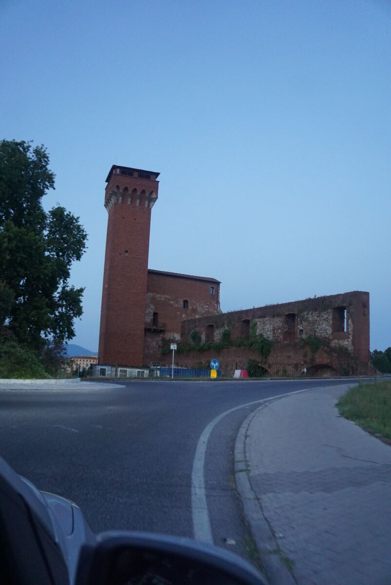 Een hoge, bakstenen toren met kantelen aan de bovenzijde staat centraal, als onderdeel van een groter, deels vervallen bakstenen bouwwerk. De lucht is helderblauw, passend bij de schemering. Op de voorgrond is een bochtige weg te zien, met aan de linkerkant bomen en rechts gras. Een deel van de auto, waaronder de spiegel, is linksonder in beeld.