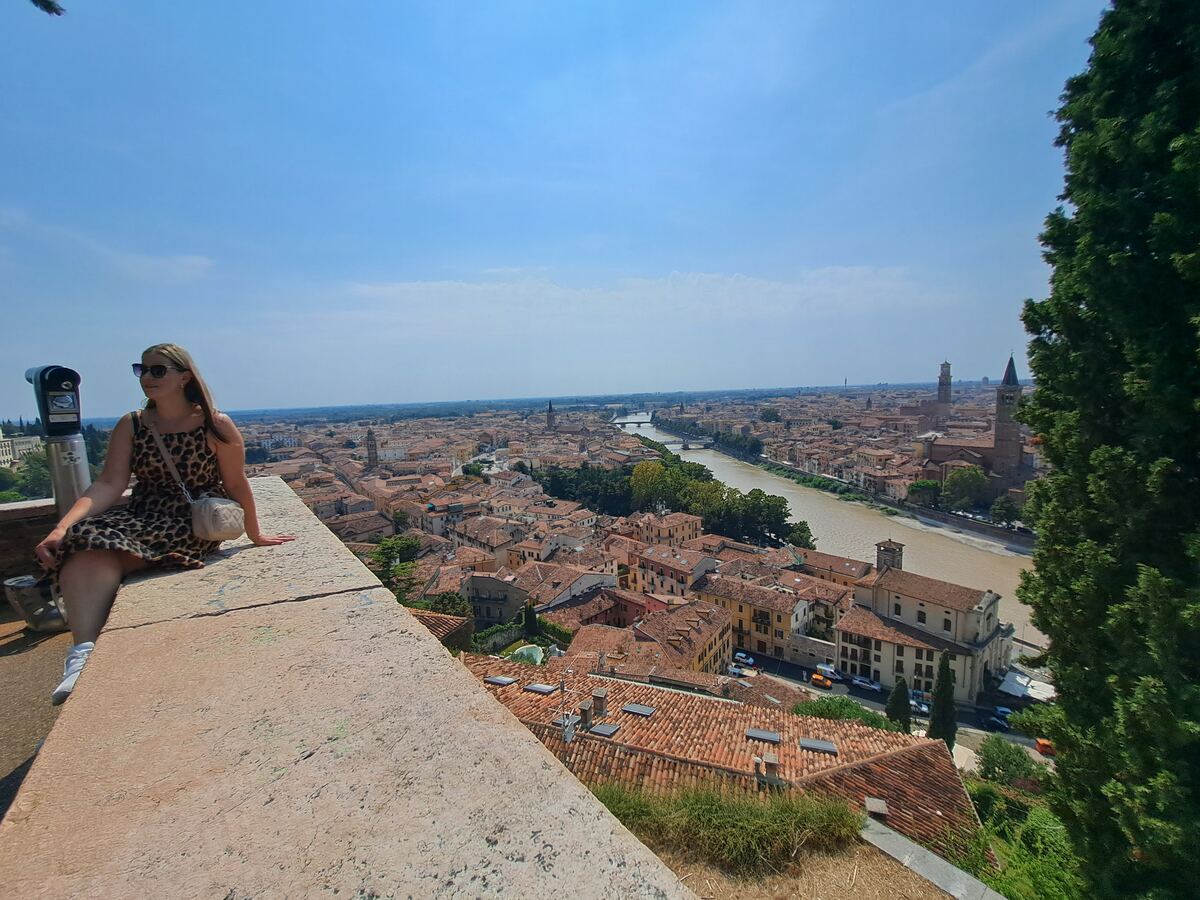 Een vrouw zit op een stenen muur met op de achtergrond een weids uitzicht over de stad Verona, met de rivier de Adige die door de stad stroomt en talloze gebouwen met oranje daken onder een helderblauwe lucht. Rechts is een deel van een cipresboom zichtbaar en links staat een verrekijker.