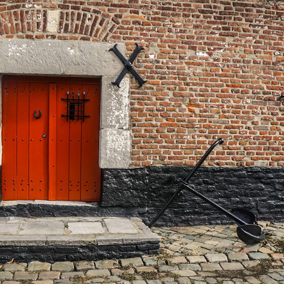 Een close-up van een bakstenen gevel met een feloranje houten deur. Boven de deur zit een stenen latei en een boog van bakstenen. Rechts van de deur is een zwart metalen kruis aan de muur bevestigd. Op de grond, rechts van de deur, ligt een groot zwart anker op een kasseienpad. De onderkant van de muur is zwart geverfd.