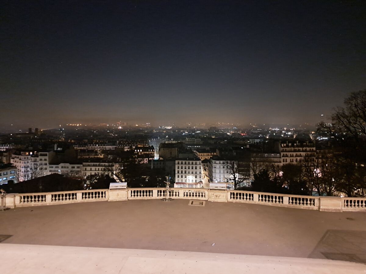 Een nachtelijke foto toont een weids uitzicht over een stad met talloze lichtjes die de gebouwen en straten verlichten. Op de voorgrond is een stenen balustrade te zien, die deel uitmaakt van een uitkijkpunt. De lucht boven de stad is donker, maar de horizon is verlicht door de gloed van de stad.