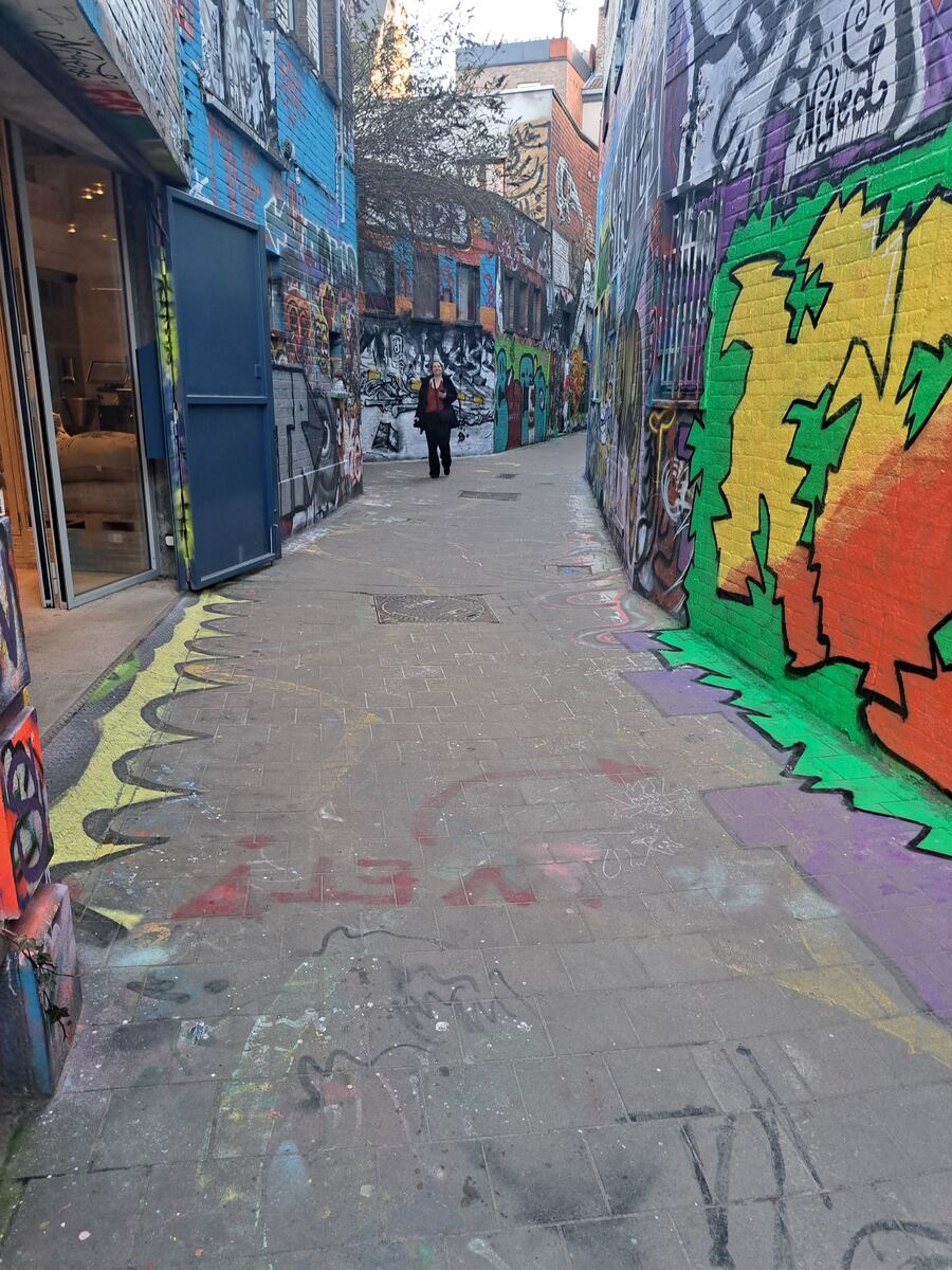 Een smalle steeg met aan beide zijden muren die volledig bedekt zijn met kleurrijke graffiti. De bestrating is van grijze tegels met hier en daar ook graffiti. Aan de linkerkant is een openstaande blauwe deur te zien en een glazen pui van een gebouw. Een persoon loopt in de verte door de steeg.