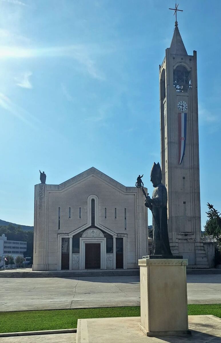 Een foto van een moderne kerk met een hoge klokkentoren ernaast, onder een helderblauwe lucht. Voor de kerk staat een standbeeld van een geestelijke en aan de klokkentoren hangt een vlag met de kleuren rood, wit en blauw. Op de achtergrond zijn heuvels te zien.