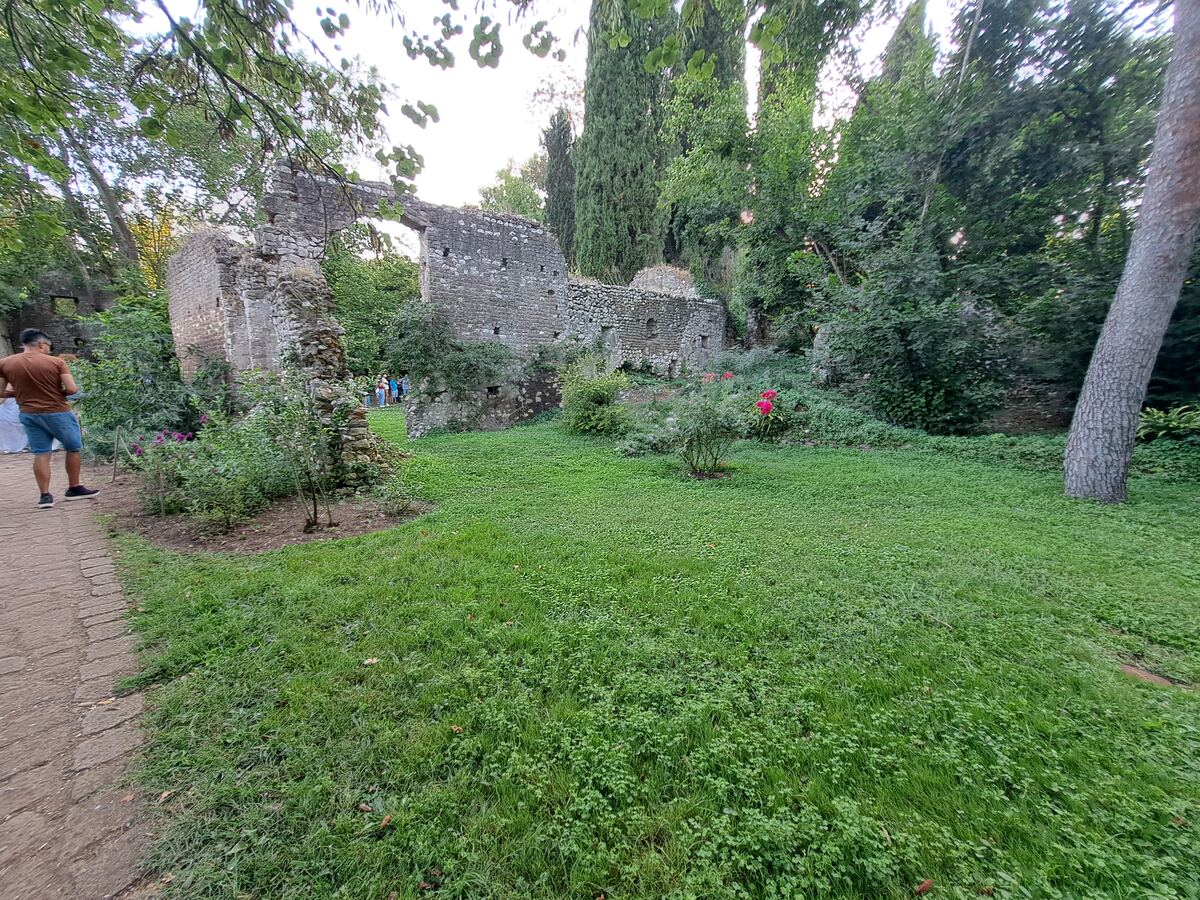 De foto toont een weids uitzicht op oude stenen ruïnes, omgeven door weelderige groene vegetatie. Centraal in beeld bevindt zich een grasveld met daarop enkele struiken, waaronder een met opvallende roze bloemen. Aan de linkerkant loopt een geplaveid pad, waarop een persoon met een bruin shirt en blauwe korte broek van de kijker wegloopt. De stenen muren van de ruïnes zijn deels ingestort en overgroeid met planten, en er zijn openingen zichtbaar die mogelijk bogen of vensters waren. Op de achter