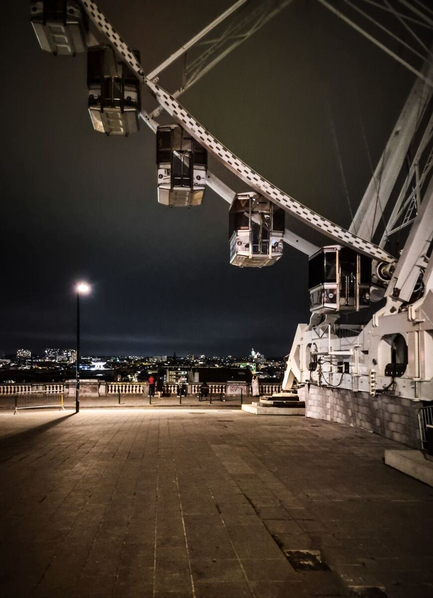 Een avondfoto van een reuzenrad met de stad op de achtergrond. De gondels van het reuzenrad zijn zichtbaar tegen de donkere lucht. Op de voorgrond is een verhard plein met een lantaarnpaal en een balustrade. Enkele mensen staan bij de balustrade en kijken naar de stad.