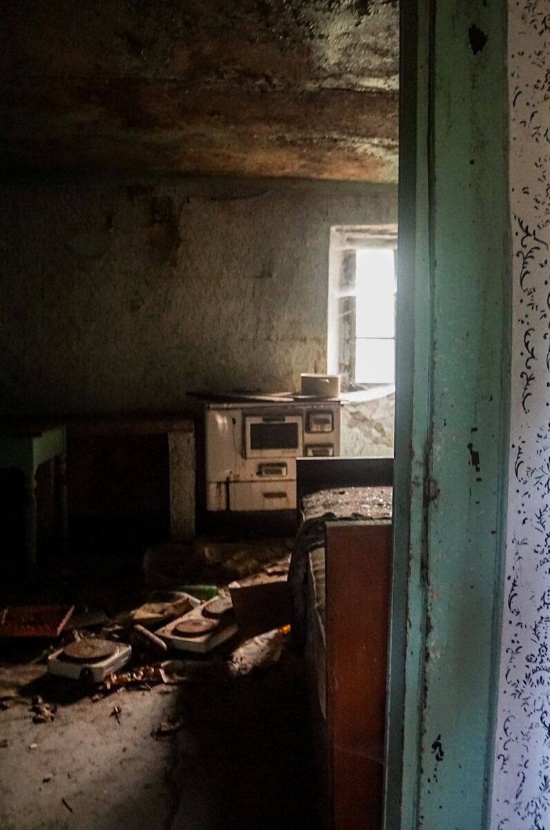 Een donkere, verlaten keuken met een oud fornuis en rommel op de vloer. Licht valt door een raam aan de achterkant. De muren zijn verweerd en het plafond is beschadigd. Rechts is een deel van een deurpost en behang met een patroon zichtbaar.