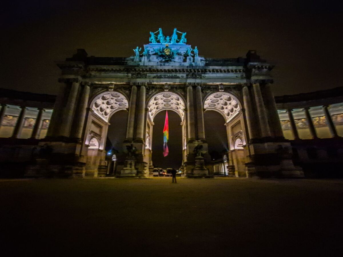 Een nachtfoto van een grote triomfboog met drie bogen, verlicht van onderaf. Bovenop de boog staan sculpturen. In de middelste boog hangt een regenboogvlag. Aan weerszijden van de boog zijn gebouwen zichtbaar met zuilen en ramen. De lucht is donker en er staat een persoon in het midden van de boog.
