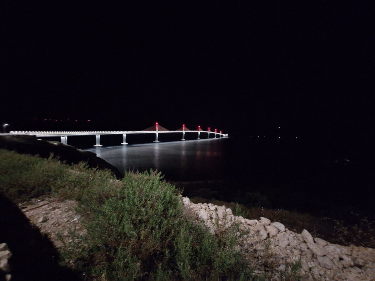 Een nachtopname van een lange brug die over water loopt, verlicht met witte en rode lichten. De brug heeft meerdere pilaren en tuien die rood oplichten. Op de voorgrond is een donkere oever met struiken en rotsen zichtbaar.