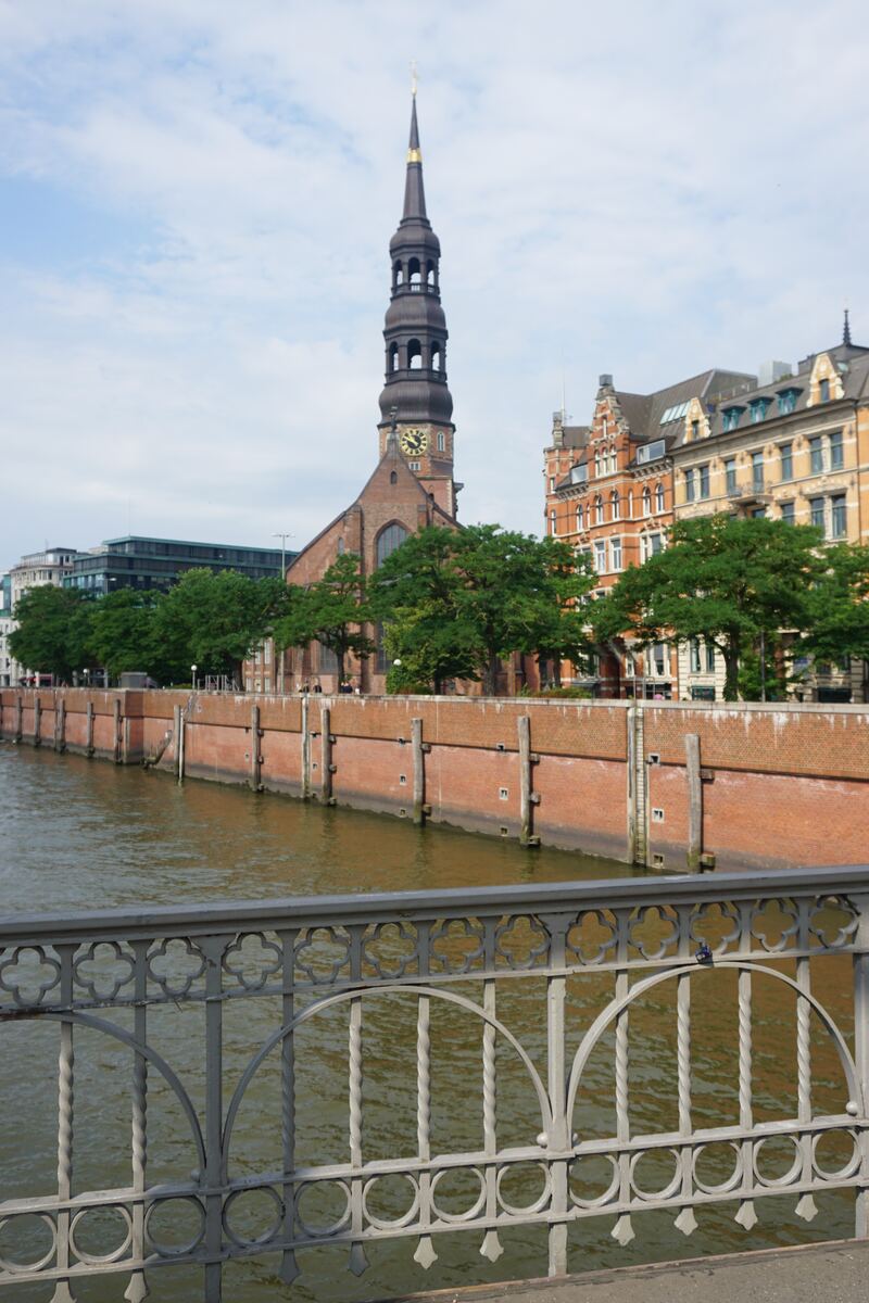 Een foto genomen vanaf een brug met een sierlijk metalen hek op de voorgrond. Op de achtergrond is een brede gracht of rivier te zien met aan de overkant een bakstenen kade. Daarachter staat de St. Catharinakerk met zijn karakteristieke toren en klok, omringd door bomen en klassieke gebouwen.