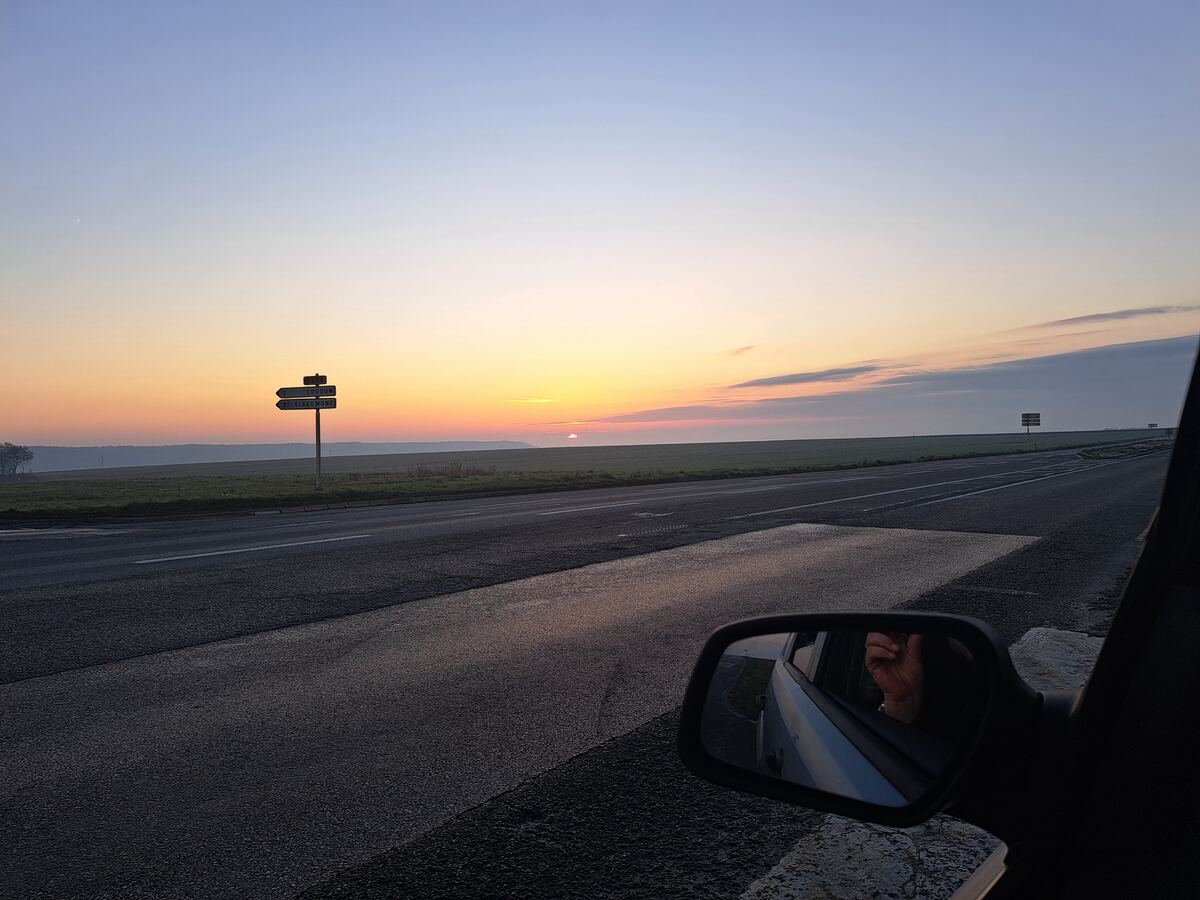 Een foto genomen vanuit een auto, met de zonsopkomst aan de horizon boven uitgestrekte groene velden. De lucht toont een kleurovergang van blauw naar oranje en roze. Links in beeld staat een wegwijzer met de namen 'Coudun' en 'Ribécourt'. De weg loopt diagonaal door het beeld en in de rechterbuitenspiegel is een deel van de auto en een hand zichtbaar.