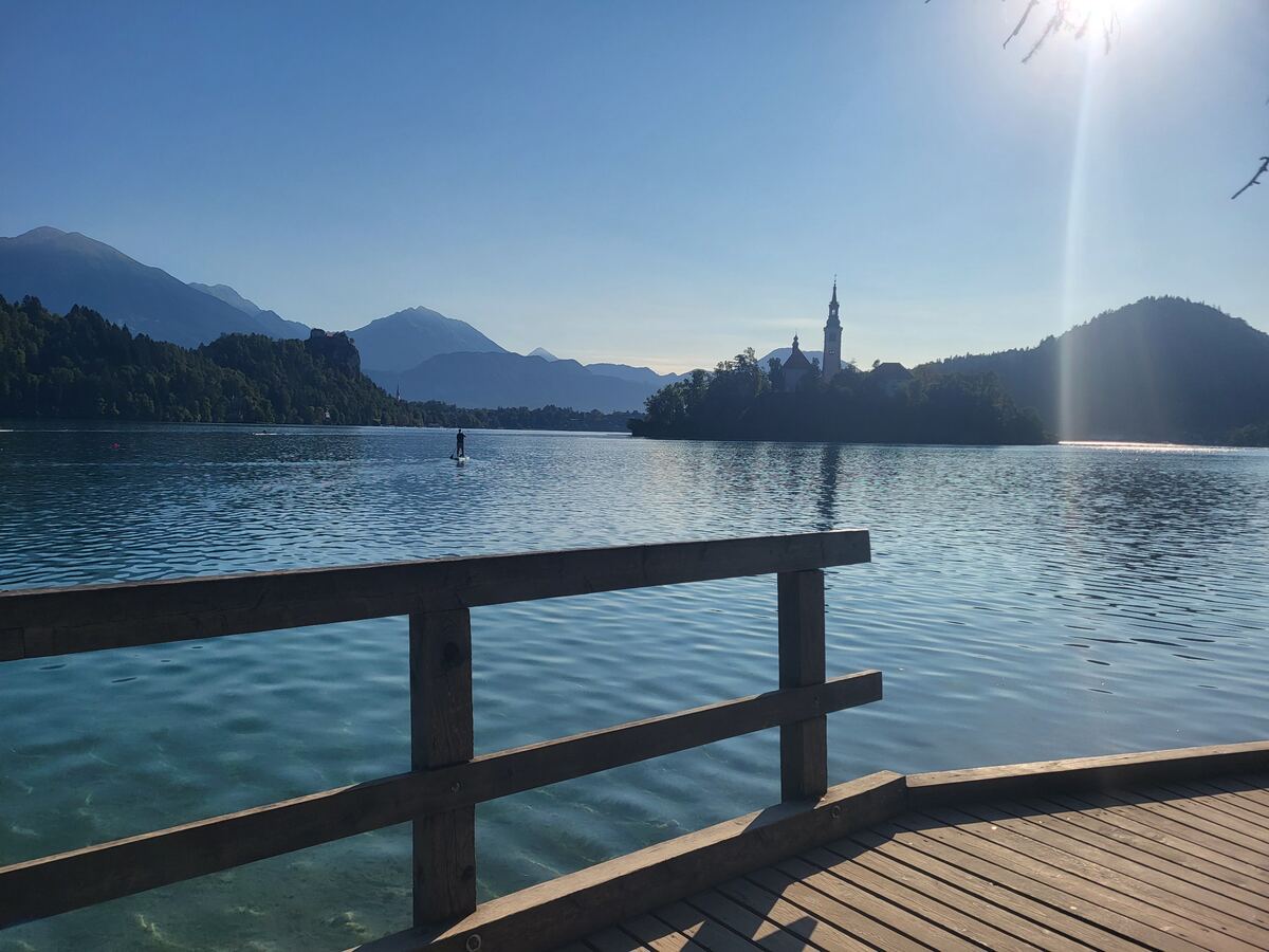 Een zonnige ochtend aan een meer met een houten steiger op de voorgrond. Op het meer is een persoon te zien op een paddleboard. In de verte ligt een eiland met een kerk en daarachter zijn bergen zichtbaar. De zon schijnt fel en reflecteert op het water.