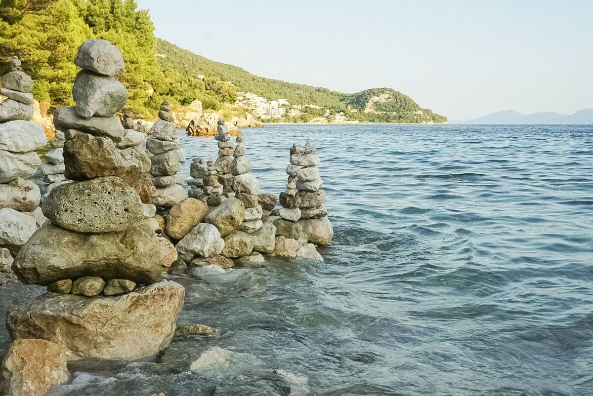 Een kustlijn met kalm, blauw water op de voorgrond en een rij gestapelde stenen, of 'cairns', die deels in het water staan. Op de achtergrond is een groene, beboste heuvel te zien met enkele witte gebouwen en in de verte liggen bergen aan de horizon.