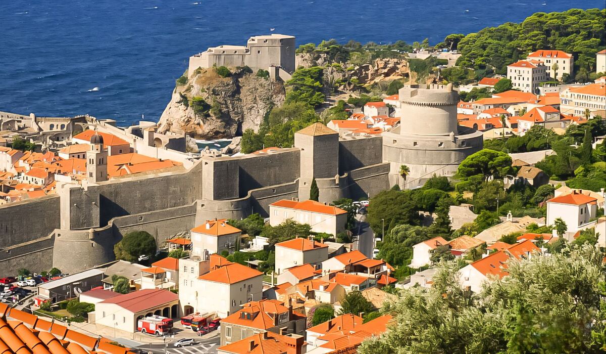 Een panoramisch uitzicht op de oude stadsmuren van Dubrovnik, met de Adriatische Zee op de achtergrond. De stadsmuren zijn van steen en omringen gebouwen met oranje daken. Er zijn ook groene bomen en struiken zichtbaar, en een deel van een weg met enkele voertuigen.
