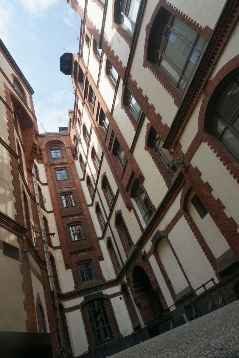 Een opwaartse foto van de gevels van meerdere hoge, historische gebouwen met rode bakstenen en crèmekleurige muren, kenmerkend voor de pakhuizen in Hamburg. De ramen zijn rechthoekig en boogvormig, en de lucht is lichtblauw met enkele wolken.