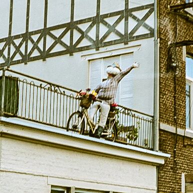 Een pop op een fiets staat op een balkon van een gebouw. De pop draagt een gestreept overhemd en een hoed, met één arm omhoog gestoken. Het gebouw heeft een gevel met vakwerk en een raam met luiken. De pop is versierd met bloemen.