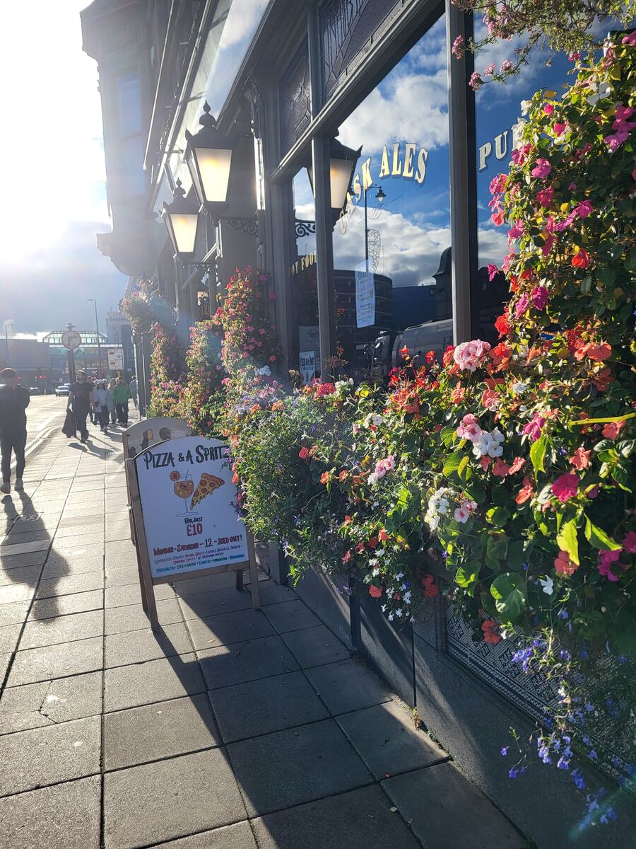 Een zonnige straatscène met een gebouw versierd met bloembakken vol kleurrijke bloemen. Op de ramen van het gebouw staat 'CASK ALES PUB' gespiegeld. Op de stoep staat een reclamebord voor 'Pizza & A Spritz' voor £10. Mensen lopen op de achtergrond en de zon schijnt fel, wat lange schaduwen werpt.