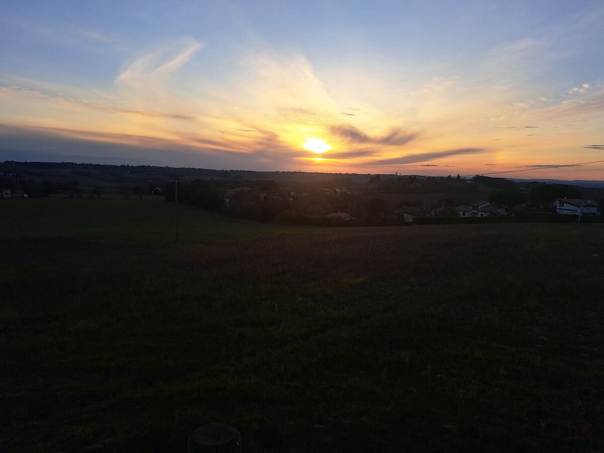 De foto toont een weids landschap bij zonsondergang. De zon staat laag aan de horizon en kleurt de lucht in tinten van geel, oranje en blauw, met enkele verspreide wolken. Op de voorgrond zijn donkere velden te zien, die overgaan in een strook bomen en een aantal verspreide huizen. Op de achtergrond strekken glooiende heuvels zich uit onder de horizon.