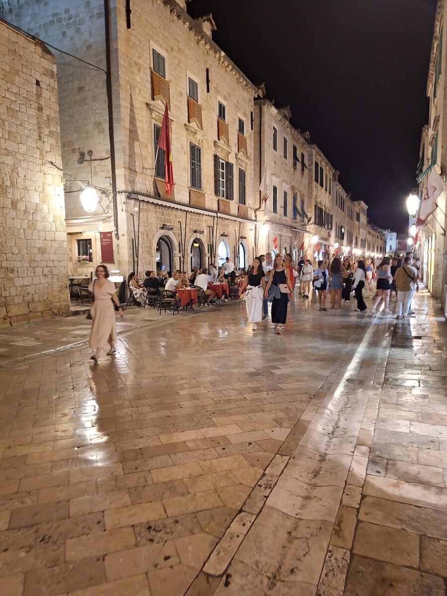 Een avondbeeld van een drukke, verlichte straat in een oude stad, met veel mensen die wandelen en enkele terrassen met gasten. De gebouwen langs de straat zijn van lichtgekleurde steen en hebben ramen met luiken. De straat zelf is geplaveid met glimmende stenen, wat wijst op natte ondergrond of reflectie van de verlichting. Er hangen vlaggen aan de gebouwen.