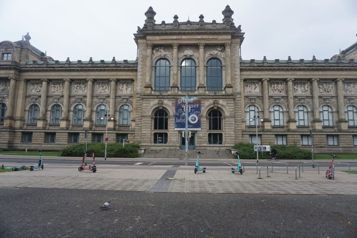 Een imposant stenen gebouw met klassieke architectuur en een banner met 'grundwasser lebt' hangend boven de ingang. Op de voorgrond staan meerdere elektrische scooters en een duif loopt over de stoep.