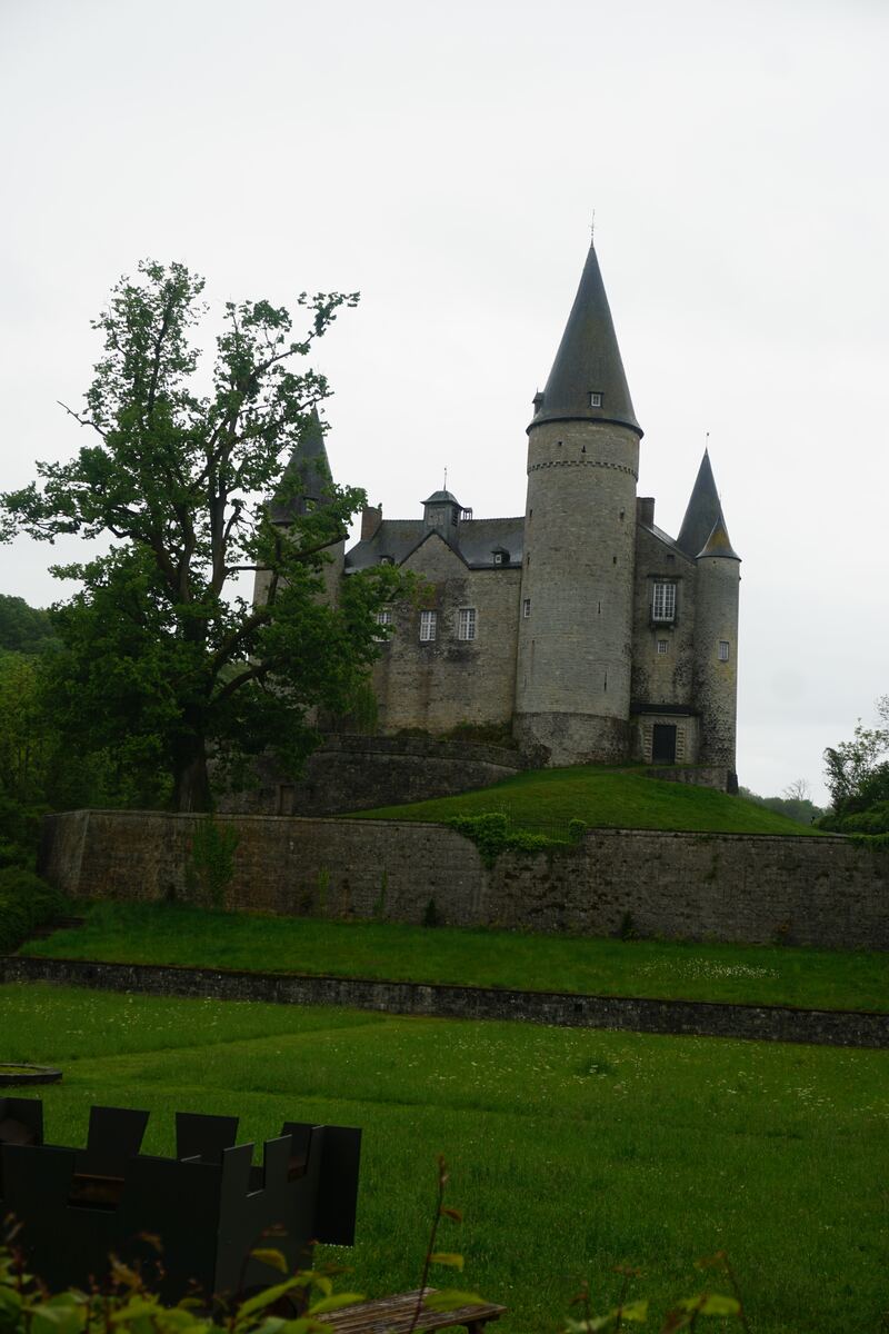 Een stenen kasteel met meerdere torens en puntige daken staat op een heuvel, omringd door groen gras en bomen onder een bewolkte hemel. Op de voorgrond is een deel van een houten bankje en een donker metalen object zichtbaar.