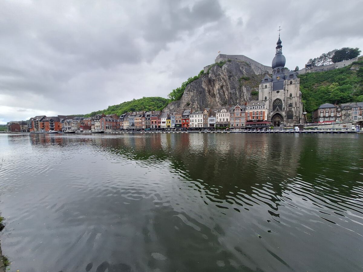 Een breed overzicht van de stad Dinant, gezien vanaf het water, met de Maas op de voorgrond. Langs de oever staan kleurrijke gebouwen, met op de achtergrond een steile rotswand en de Citadel van Dinant. De Collegiale kerk Onze-Lieve-Vrouw van Dinant met zijn opvallende uivormige koepel is duidelijk zichtbaar.