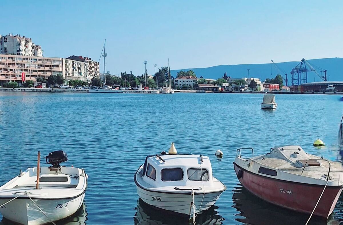 Een zonnige foto van de haven van Ploče met op de voorgrond drie kleine boten in het water. Op de achtergrond zijn gebouwen, bomen en een deel van de stad te zien, met daarachter bergen en een helderblauwe lucht. Rechts in de verte zijn havenkranen zichtbaar.